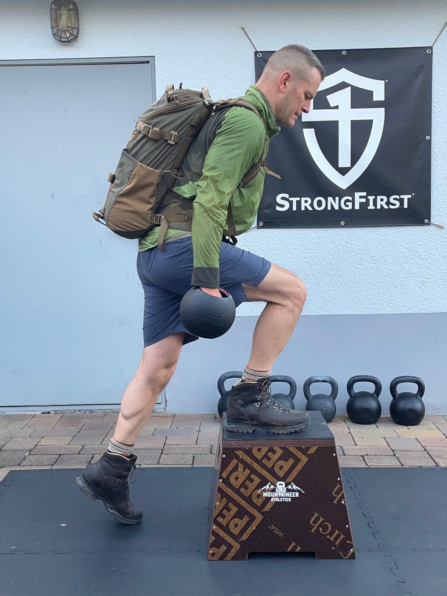 Kenneth Bolyard mit Wanderschuhen und beladenem Rucksack steigt im Gym mit einer Kettlebell in der Hand auf eine Box